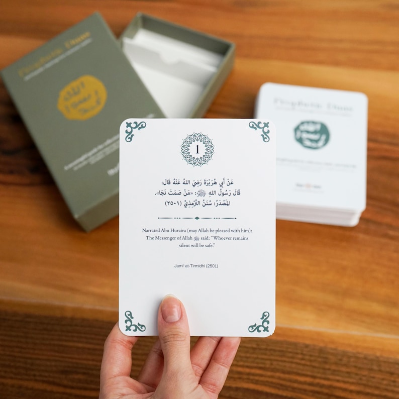 K&ouml;nnte beinhalten: Ein Satz wei&szlig;er Karten mit arabischem Text und englischer &Uuml;bersetzung, gehalten von einer Hand. Die Karten befinden sich in einer Schachtel mit gr&uuml;nem Deckel. Der Text enth&auml;lt ein Zitat des Gesandten Allahs. Die Karten liegen auf einer Holzoberfl&auml;che.