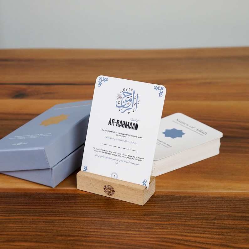 K&ouml;nnte beinhalten: Ein Satz wei&szlig;er Karten mit blauem Text und Designs, der auf einem Holzst&auml;nder pr&auml;sentiert wird. Die Karten sind mit "AR-RAHMAAN" und "Namen Allahs" beschriftet. Eine hellblaue Box mit einem goldenen Design ist ebenfalls sichtbar.