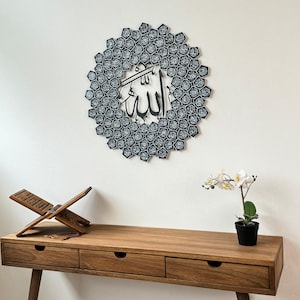 Peut inclure: Une d&eacute;coration murale noire et blanche repr&eacute;sentant le mot arabe "Allah" entour&eacute; d'un motif circulaire de petits carr&eacute;s noirs et blancs. La d&eacute;coration est accroch&eacute;e &agrave; un mur blanc au-dessus d'une table en bois avec un pupitre de pri&egrave;re en bois et une fleur blanche dans un pot.