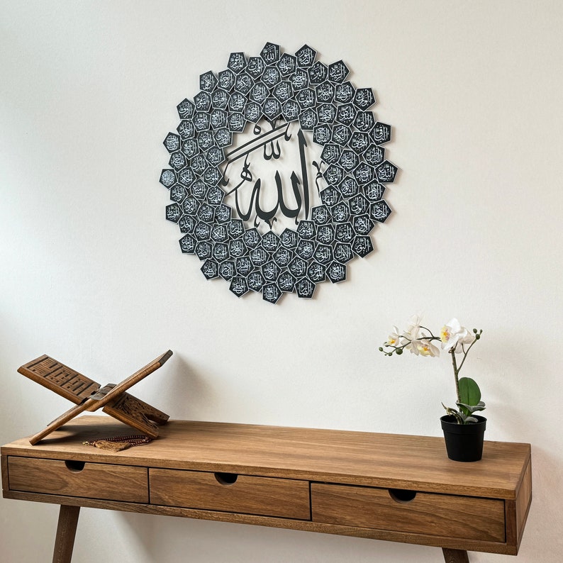 Peut inclure: Une d&eacute;coration murale noire et blanche repr&eacute;sentant le mot arabe "Allah" entour&eacute; d'un motif circulaire de petits carr&eacute;s noirs et blancs. La d&eacute;coration est accroch&eacute;e &agrave; un mur blanc au-dessus d'une table en bois avec un pupitre de pri&egrave;re en bois et une fleur blanche dans un pot.