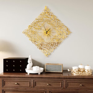 May include: A gold-colored, diamond-shaped wall clock with intricate Arabic calligraphy. The clock has a sunburst design around the clock hands. The clock is mounted on a light gray wall above a wooden dresser with decorative items.