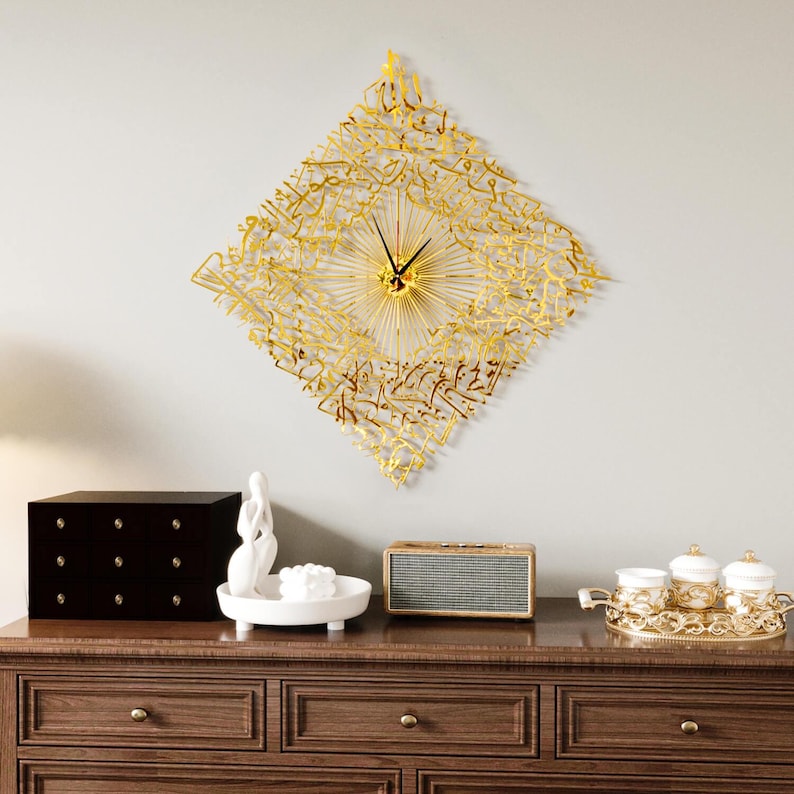 May include: A gold-colored, diamond-shaped wall clock with intricate Arabic calligraphy. The clock has a sunburst design around the clock hands. The clock is mounted on a light gray wall above a wooden dresser with decorative items.