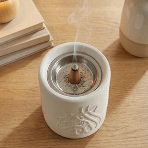 Peut inclure: Un porte-encens en c&eacute;ramique blanche avec un insert en m&eacute;tal, contenant un c&ocirc;ne d'encens en combustion. Le support pr&eacute;sente un motif en relief sur le c&ocirc;t&eacute;. De la fum&eacute;e s'&eacute;l&egrave;ve du c&ocirc;ne, cr&eacute;ant une ambiance paisible.