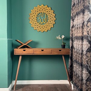 Peut inclure: Un bureau en bois avec trois tiroirs et une d&eacute;coration murale en m&eacute;tal dor&eacute; avec le mot "Allah" en calligraphie arabe. Le bureau est devant un mur vert sarcelle et un rideau &agrave; motifs.