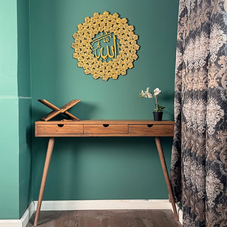 Peut inclure: Un bureau en bois avec trois tiroirs et une d&eacute;coration murale en m&eacute;tal dor&eacute; avec le mot "Allah" en calligraphie arabe. Le bureau est devant un mur vert sarcelle et un rideau &agrave; motifs.