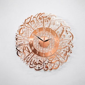 May include: A copper-colored wall clock with Arabic calligraphy surrounding the clock face. The clock face has black hands and black Roman numeral markings.