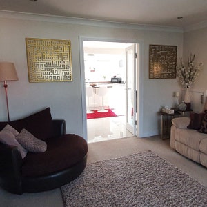 May include: A living room with a brown leather swivel chair, a beige rug, and two gold metal wall decorations with Arabic calligraphy. The room has a doorway leading to a kitchen with white cabinets and a red rug.
