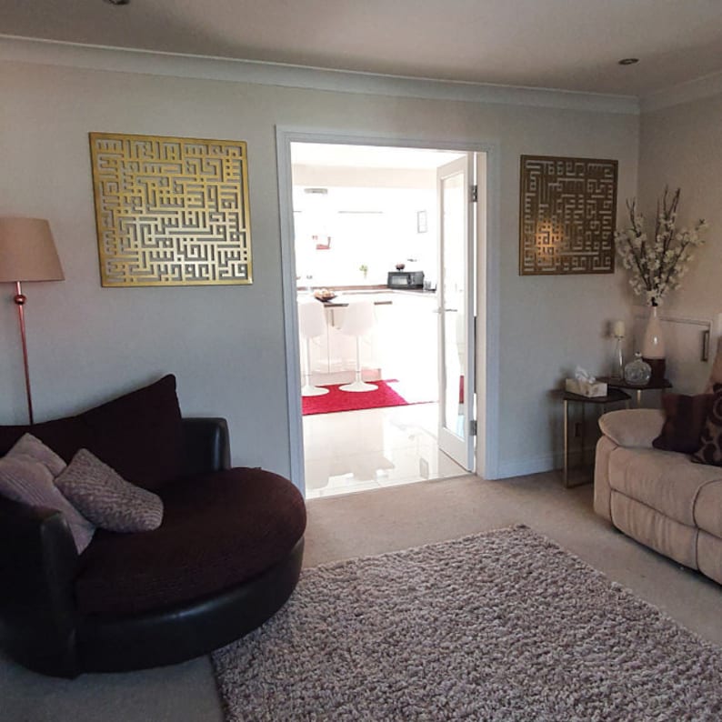 May include: A living room with a brown leather swivel chair, a beige rug, and two gold metal wall decorations with Arabic calligraphy. The room has a doorway leading to a kitchen with white cabinets and a red rug.