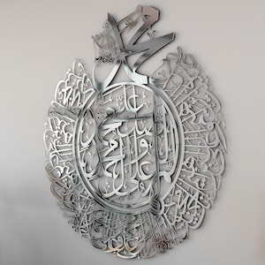 May include: A silver metal wall art featuring intricate Arabic calligraphy in a circular design. The calligraphy is in a traditional style and is surrounded by a decorative border.