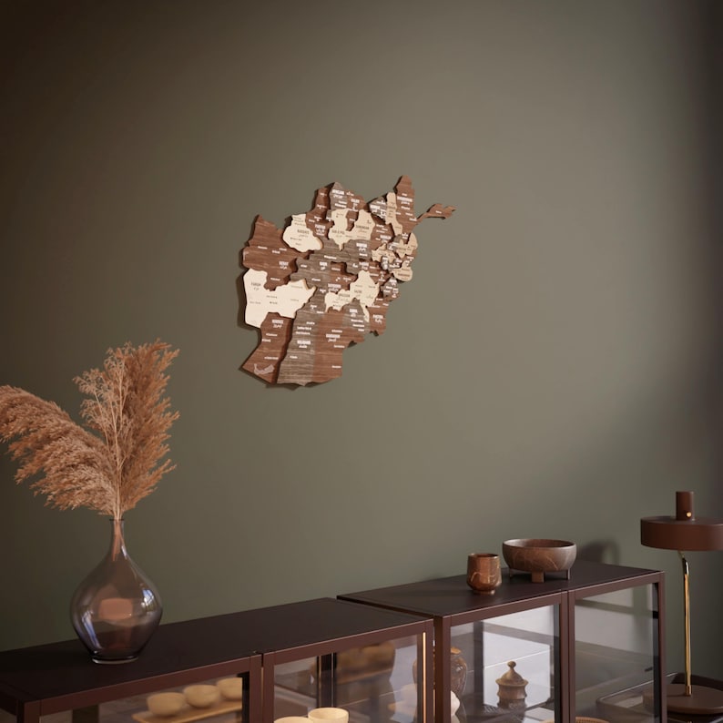 May include: A wooden map of Afghanistan with the names of each province labeled on the map. The map is hanging on a green wall with a brown wooden shelf below it. The shelf has a vase with dried flowers, a small bowl, and a larger bowl.
