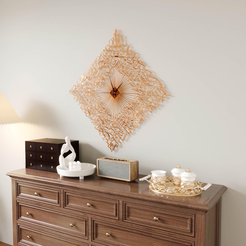 May include: A gold-colored, diamond-shaped wall clock with Arabic calligraphy. The clock has a radial design with a red center and hands. The clock hangs above a wooden dresser with decorative items, including a speaker and a tray with cups.