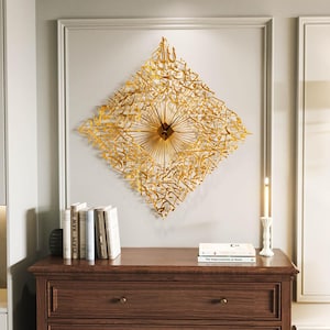 May include: A gold-colored, diamond-shaped wall clock with intricate Arabic calligraphy. The clock features a central clock face with radiating lines and black hands. The clock is mounted on a light gray wall above a wooden dresser.