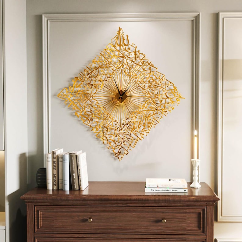 May include: A gold-colored, diamond-shaped wall clock with intricate Arabic calligraphy. The clock features a central clock face with radiating lines and black hands. The clock is mounted on a light gray wall above a wooden dresser.