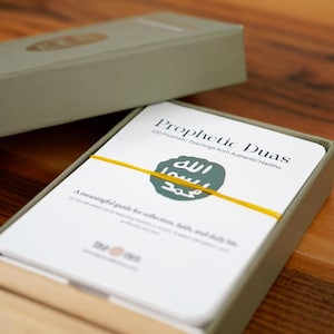 K&ouml;nnte beinhalten: Ein Satz Prophetic Duas-Karten in einer grauen Box. Die Karten sind wei&szlig; mit dem Titel "Prophetic Duas" und einem gr&uuml;nen Emblem. Ein gelbes Gummiband h&auml;lt die Karten zusammen. Die Box ist ge&ouml;ffnet und zeigt die Karten.