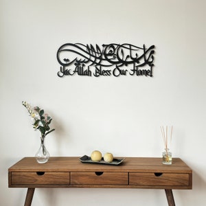 May include: Black metal wall art with Arabic calligraphy that reads "Ya Allah Bless Our Home". The art is mounted on a white wall above a wooden table with two decorative yellow spheres on a black tray.