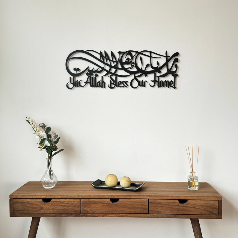 May include: Black metal wall art with Arabic calligraphy that reads "Ya Allah Bless Our Home". The art is mounted on a white wall above a wooden table with two decorative yellow spheres on a black tray.