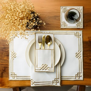 May include: Elegant white placemat, napkin, and cutlery set with gold trim and geometric designs. Includes a gold fork, knife, and spoon. A matching coaster and a glass are also present. The table setting is on a wooden surface.