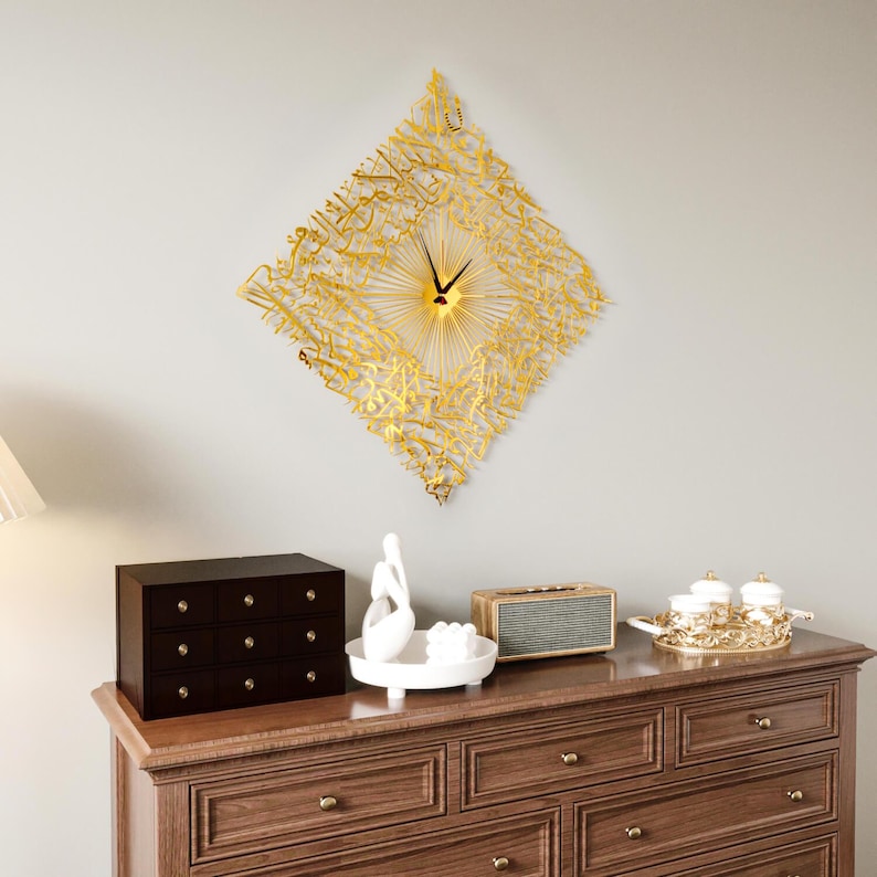 May include: A gold-colored, diamond-shaped wall clock with Arabic calligraphy. The clock has a sunburst design in the center and black clock hands. The clock is mounted on a light gray wall above a wooden dresser.