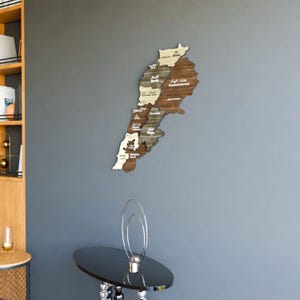 May include: A wooden map of Lebanon with the names of each region labelled in Arabic. The map is mounted on a grey wall and is surrounded by a white frame.