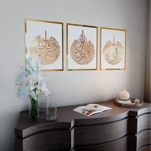 May include: Three gold framed prints with white backgrounds and gold Arabic calligraphy inside each frame. The frames are hanging on a white wall above a dark wood dresser.