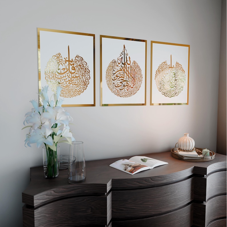 May include: Three gold framed prints with white backgrounds and gold Arabic calligraphy inside each frame. The frames are hanging on a white wall above a dark wood dresser.