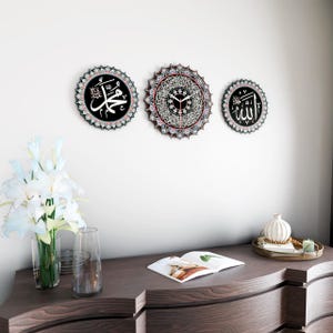 Op de afbeelding: Drie zwarte ronde wanddecoraties met ingewikkelde witte en rode ontwerpen. Het middelste stuk heeft een klok met Arabische cijfers. Het linker stuk heeft het Arabische woord "Muhammad" in witte letters. Het rechter stuk heeft het Arabische woord "Allah" in witte letters.