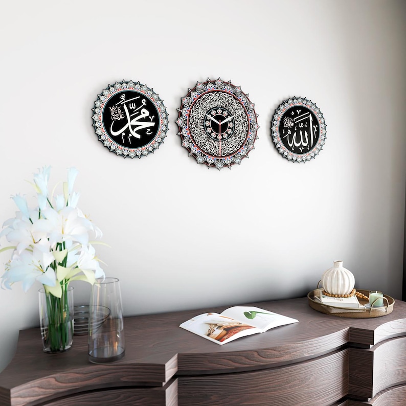 Op de afbeelding: Drie zwarte ronde wanddecoraties met ingewikkelde witte en rode ontwerpen. Het middelste stuk heeft een klok met Arabische cijfers. Het linker stuk heeft het Arabische woord "Muhammad" in witte letters. Het rechter stuk heeft het Arabische woord "Allah" in witte letters.