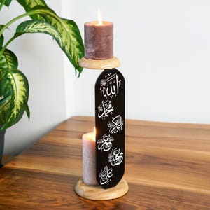 May include: A decorative candle holder with two lit candles. The holder is black with white Arabic calligraphy. The candles are a muted pink and brown. The holder is made of wood and sits on a wooden table.