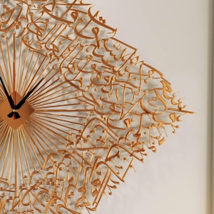 May include: Gold-colored wall clock with a unique design. The clock features a square center with radiating lines and black clock hands. Intricate gold calligraphy covers the clock face, adding an artistic touch to the decor.