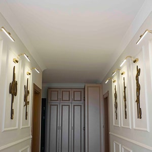 May include: A hallway with white walls and decorative molding. Gold-toned wall sconces with integrated LED lights are mounted above gold-colored wall art. The art features stylized calligraphy. The hallway leads to a light-colored cabinet.