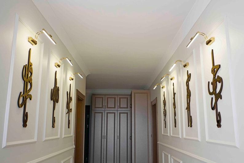 May include: A hallway with white walls and decorative molding. Gold-toned wall sconces with integrated LED lights are mounted above gold-colored wall art. The art features stylized calligraphy. The hallway leads to a light-colored cabinet.