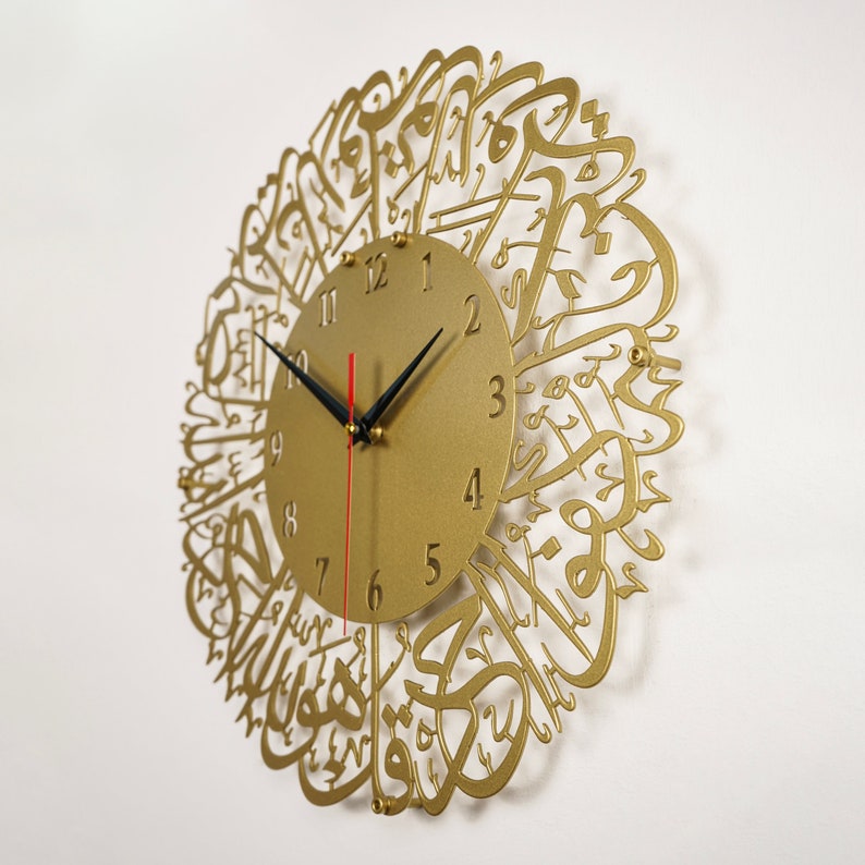 K&ouml;nnte beinhalten: Eine goldfarbene Wanduhr mit arabischer Kalligraphie, die das Zifferblatt umgibt. Das Zifferblatt hat schwarze Zeiger und schwarze Zahlen.