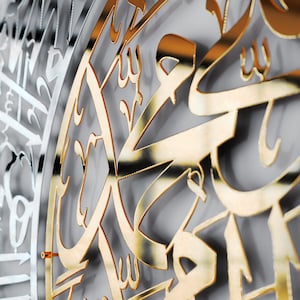 May include: A close-up of a gold-colored metal wall art piece with intricate Arabic calligraphy. The design is circular and features a repeating pattern of stylized letters.