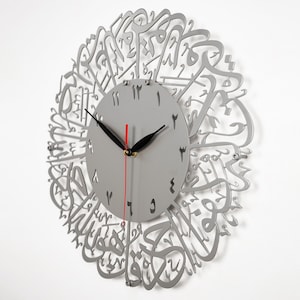 K&ouml;nnte beinhalten: Eine silberne Wanduhr mit arabischer Kalligraphie um das Zifferblatt. Das Zifferblatt der Uhr hat schwarze Zeiger und arabische Ziffern.