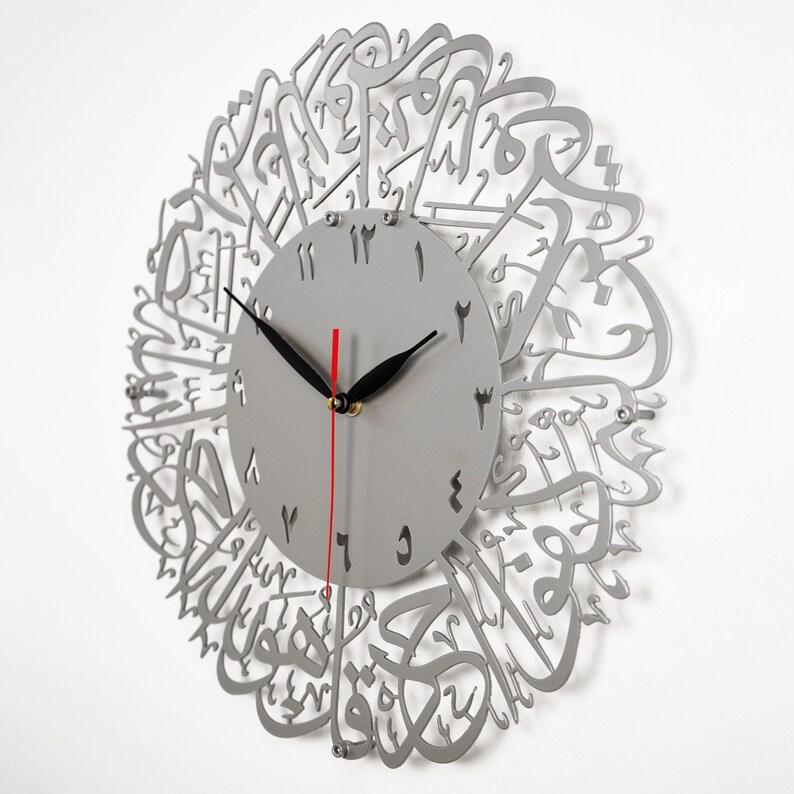 K&ouml;nnte beinhalten: Eine silberne Wanduhr mit arabischer Kalligraphie um das Zifferblatt. Das Zifferblatt der Uhr hat schwarze Zeiger und arabische Ziffern.