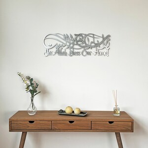 May include: A silver metal wall art with Arabic calligraphy that reads "Ya Allah Bless Our Home". The art is hanging on a white wall above a wooden table with three drawers. There are two yellow decorative spheres on a black tray on the table.