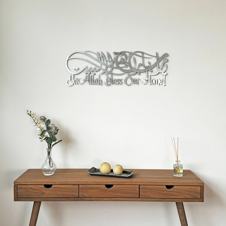 May include: A silver metal wall art with Arabic calligraphy that reads "Ya Allah Bless Our Home". The art is hanging on a white wall above a wooden table with three drawers. There are two yellow decorative spheres on a black tray on the table.