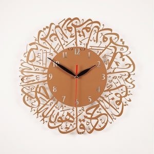 K&ouml;nnte beinhalten: Eine goldfarbene Wanduhr mit arabischer Kalligraphie, die das Zifferblatt umgibt. Das Zifferblatt ist braun mit schwarzen Zeigern und silbernen Zahlen.