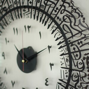 May include: A black and white clock with Arabic numerals. The clock face is white with black numbers and hands. The outer ring of the clock is made of black metal and features Arabic calligraphy.