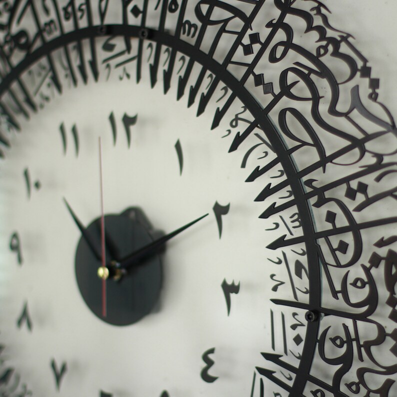 May include: A black and white clock with Arabic numerals. The clock face is white with black numbers and hands. The outer ring of the clock is made of black metal and features Arabic calligraphy.