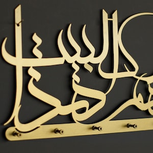 K&ouml;nnte beinhalten: Goldfarbener Schl&uuml;sselhalter aus Metall mit dem arabischen Satz "Bismillah" in einer dekorativen, stilisierten Schrift. Der Halter hat f&uuml;nf Haken zum Aufh&auml;ngen von Schl&uuml;sseln.
