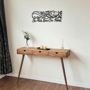 May include: A black metal wall art piece with Arabic calligraphy that reads "Ya Allah Bless Our Home". The art is hanging on a white wall above a wooden console table with three drawers. The table has a light brown finish and is sitting on a wooden floor.