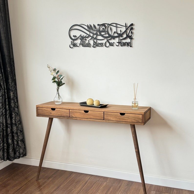 May include: A black metal wall art piece with Arabic calligraphy that reads "Ya Allah Bless Our Home". The art is hanging on a white wall above a wooden console table with three drawers. The table has a light brown finish and is sitting on a wooden floor.