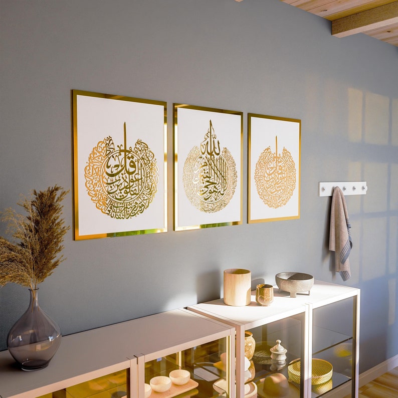 May include: Three framed gold calligraphy prints with Arabic script on a white background. The frames are gold and have a simple design. The prints are hanging on a gray wall.
