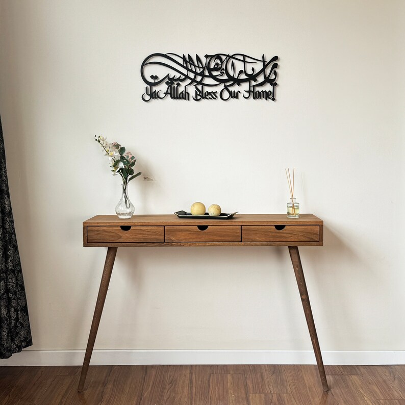 May include: A wooden console table with three drawers and tapered legs. The table is decorated with a black metal wall art featuring Arabic calligraphy that reads "Ya Allah Bless Our Home".