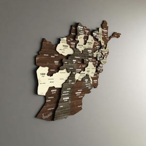 May include: A wooden map of Afghanistan with the names of each province in English and the local language. The map is made of different shades of brown wood and is mounted on a gray wall.