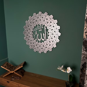 Peut inclure: Une d&eacute;coration murale en m&eacute;tal argent&eacute; repr&eacute;sentant le mot arabe "Allah" entour&eacute; d'un cercle de petits hexagones, chacun contenant un mot arabe diff&eacute;rent. La d&eacute;coration est fix&eacute;e sur un mur vert.