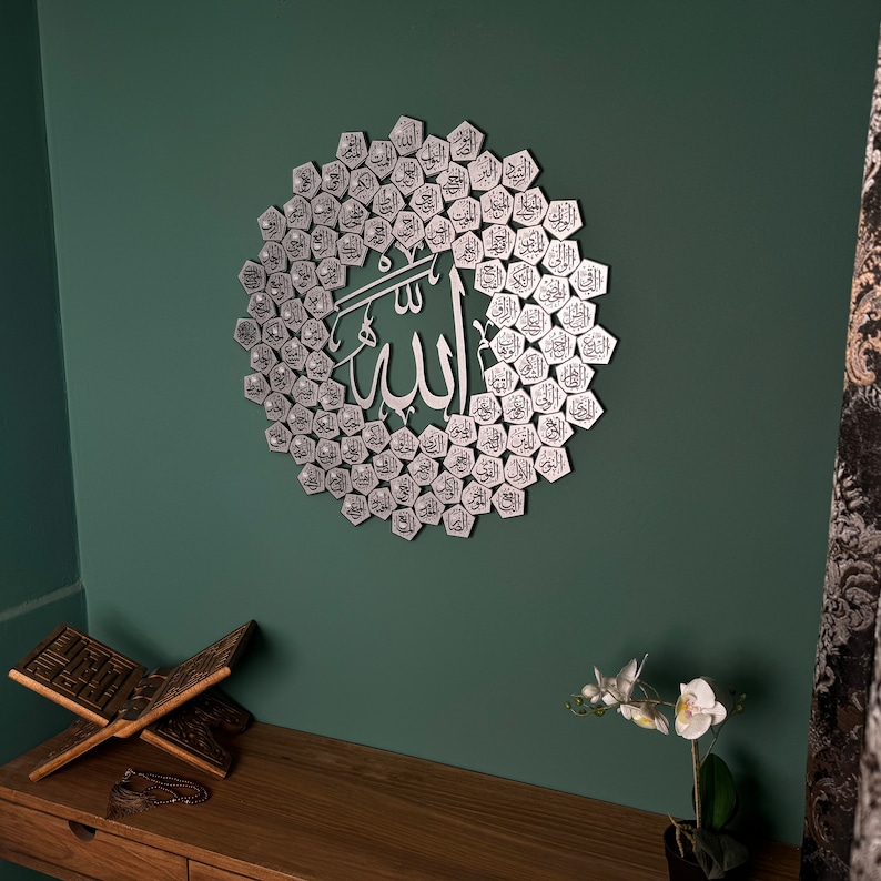 Peut inclure: Une d&eacute;coration murale en m&eacute;tal argent&eacute; repr&eacute;sentant le mot arabe "Allah" entour&eacute; d'un cercle de petits hexagones, chacun contenant un mot arabe diff&eacute;rent. La d&eacute;coration est fix&eacute;e sur un mur vert.