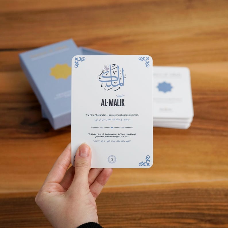 K&ouml;nnte beinhalten: Eine wei&szlig;e Karte mit dem arabischen Text "AL-MALIK" in blauer Schrift wird in einer Hand gehalten. Die Karte hat dekorative blaue Eckdesigns und Text in Englisch und Arabisch. Eine hellblaue Box mit einem goldenen Design befindet sich im Hintergrund.