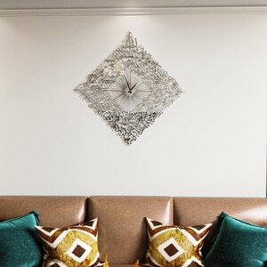 May include: A silver-colored, diamond-shaped wall clock with intricate calligraphy design. The clock features a radial design emanating from the center, with black clock hands. The clock is mounted on a white wall above a brown sofa with decorative pillows.